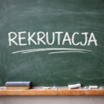 Zdjęcie przedstawia zieloną tablicę szkolną z wyraźnym napisem „REKRUTACJA” wykonanym białą kredą dużymi literami. Pod napisem znajduje się kredowa, falista linia. Na drewnianej półce u dołu tablicy leżą kawałki białej kredy oraz szkolna gąbka do ścierania, a na powierzchni tablicy widoczne są ślady i pył kredowy.